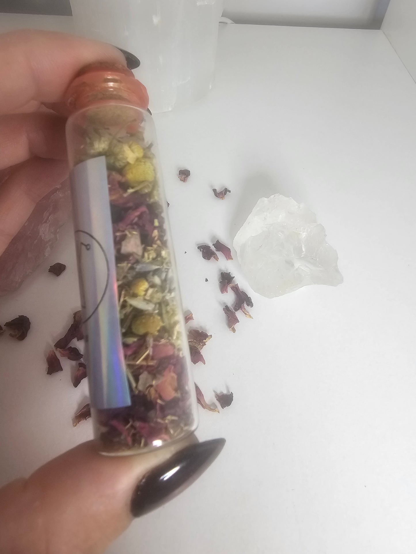 A hand with black glass nails holding a small glass spell jar filled with herbs of different colours and crystals.