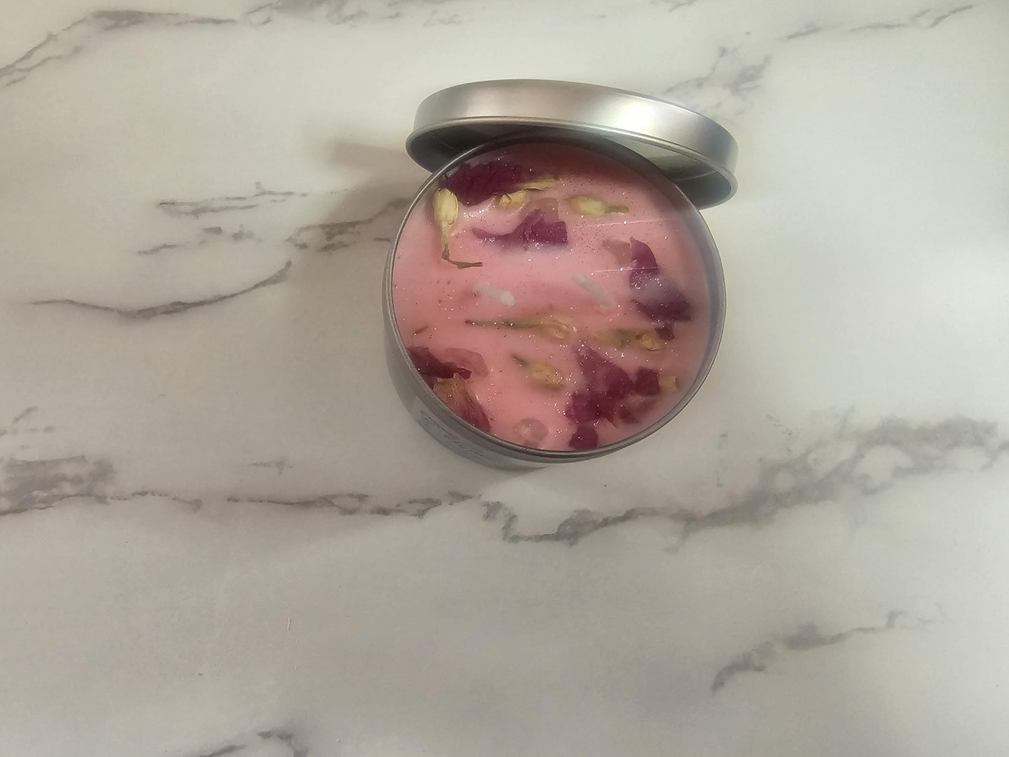 An above shot of a silver candle container filled with light pink wax with a lid resting aside the candle on a white marble background. The candle shimmers with pink and white glitter and is decorated with pink lotus petals and white jasmine buds.