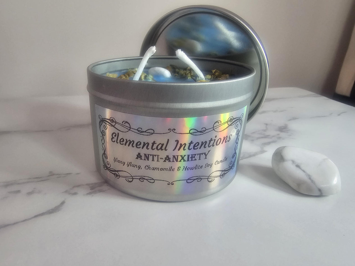 A silver container candle with a silver lid behind it on a white marble surface, next to a large howlite crystal. The holographic silver label is framed with a fine lined floral looking frame and the words on it say 'Elemental Intentions', 'anti-anxiety', and  'Ylang ylang, chamomile and howlite soy wax candle'.