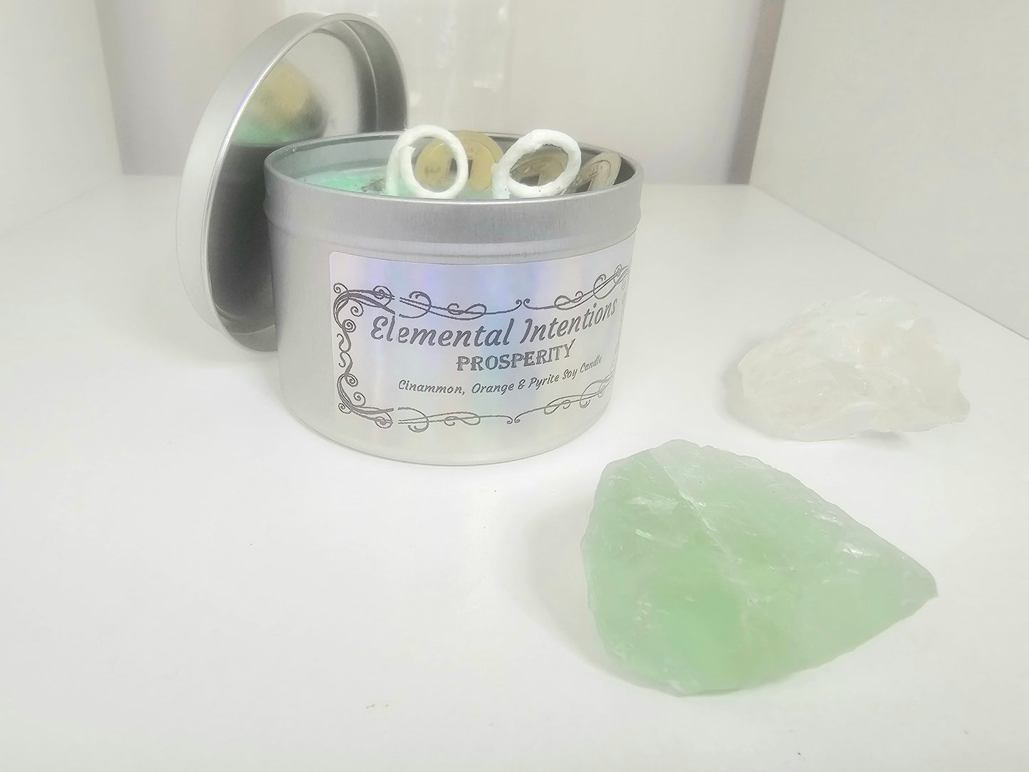 Candle in a tin labeled 'Elemental Intentions Prosperity' with a green crystal on a white surface.