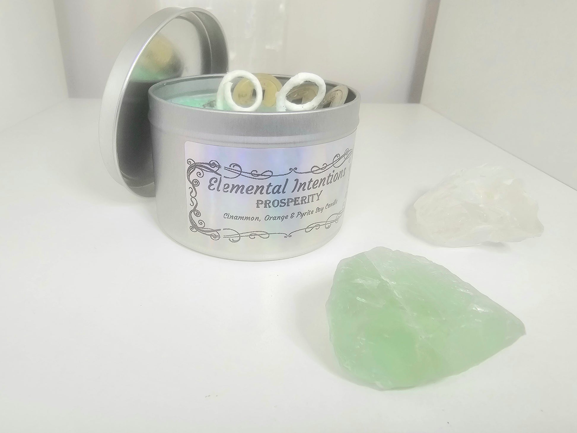 Candle in a tin labeled 'Elemental Intentions Prosperity' with a green crystal on a white surface.