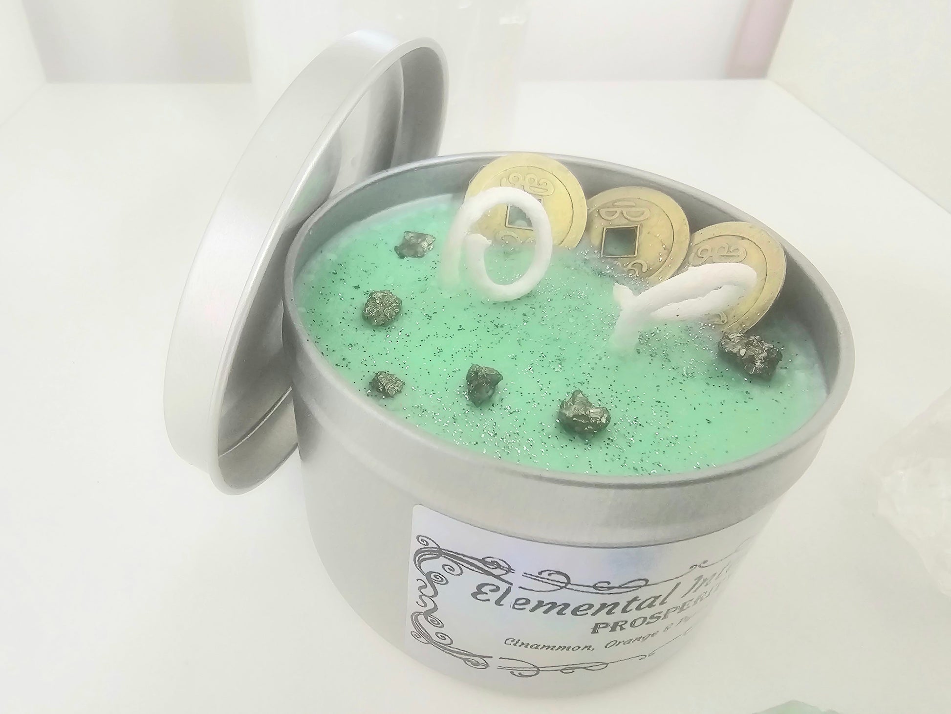 Silver candle tin filled with green scented soy wax and topped with feng shui coins, silver glitter and silver pyrite crystals.
