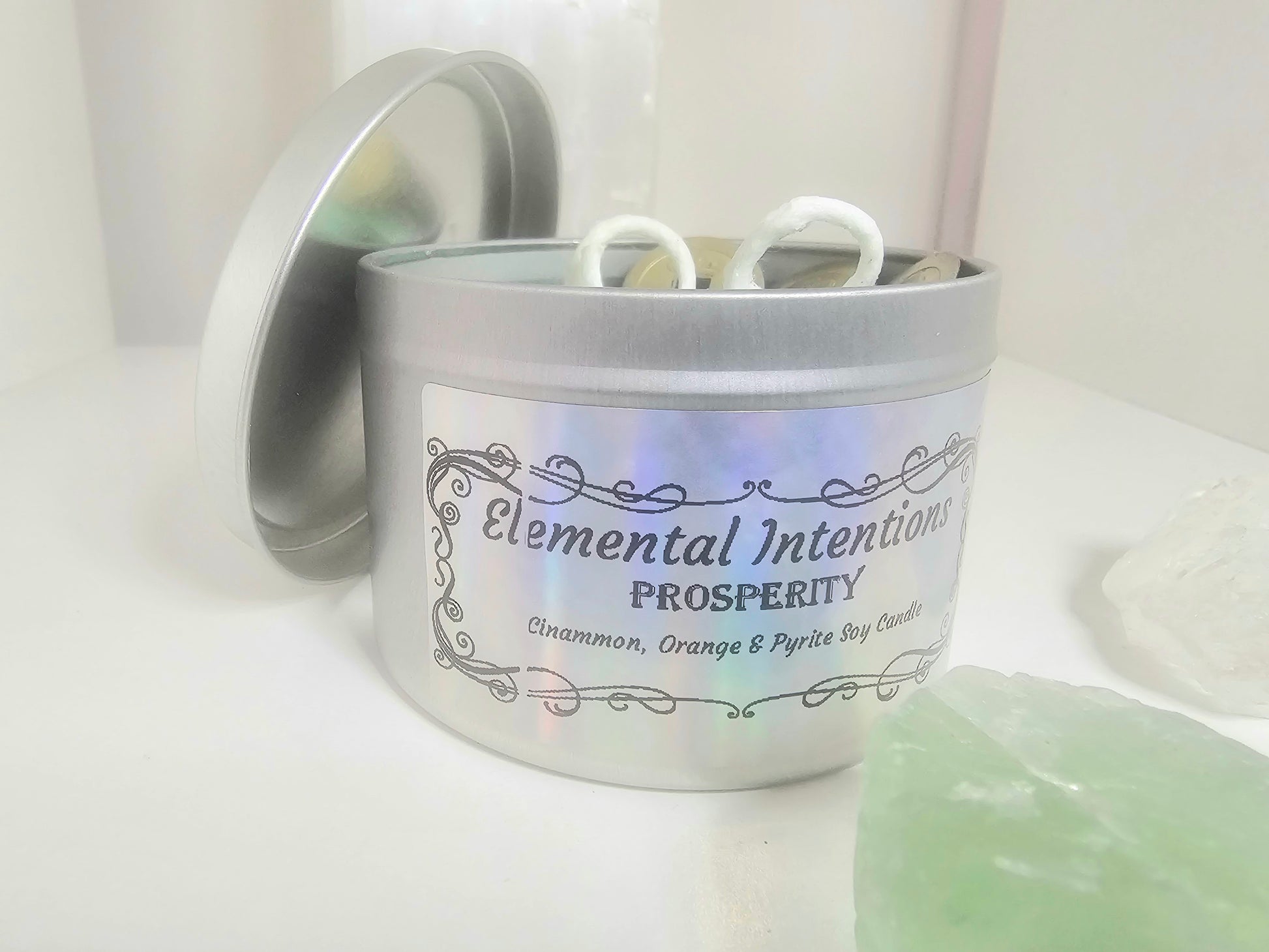 Front shot of a silver candle container with a silver holographic label that says 'elemental intentions, 'prosperity' and 'cinnamon, orange and pyrite soy candle' on a white background.