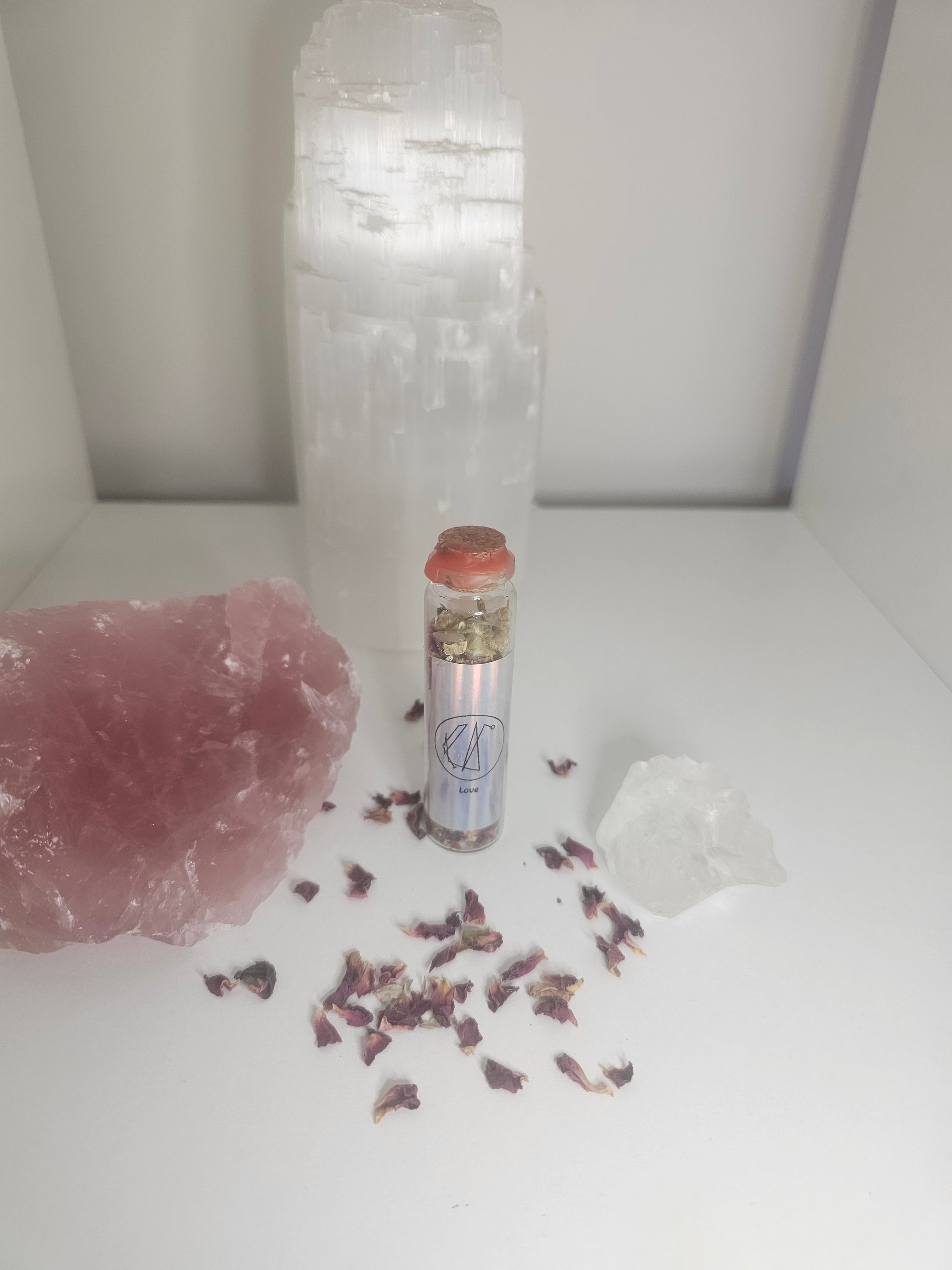 Small spell jar filled with crystals and herbs on a white background and surrounded by rose quartz and rose petals.