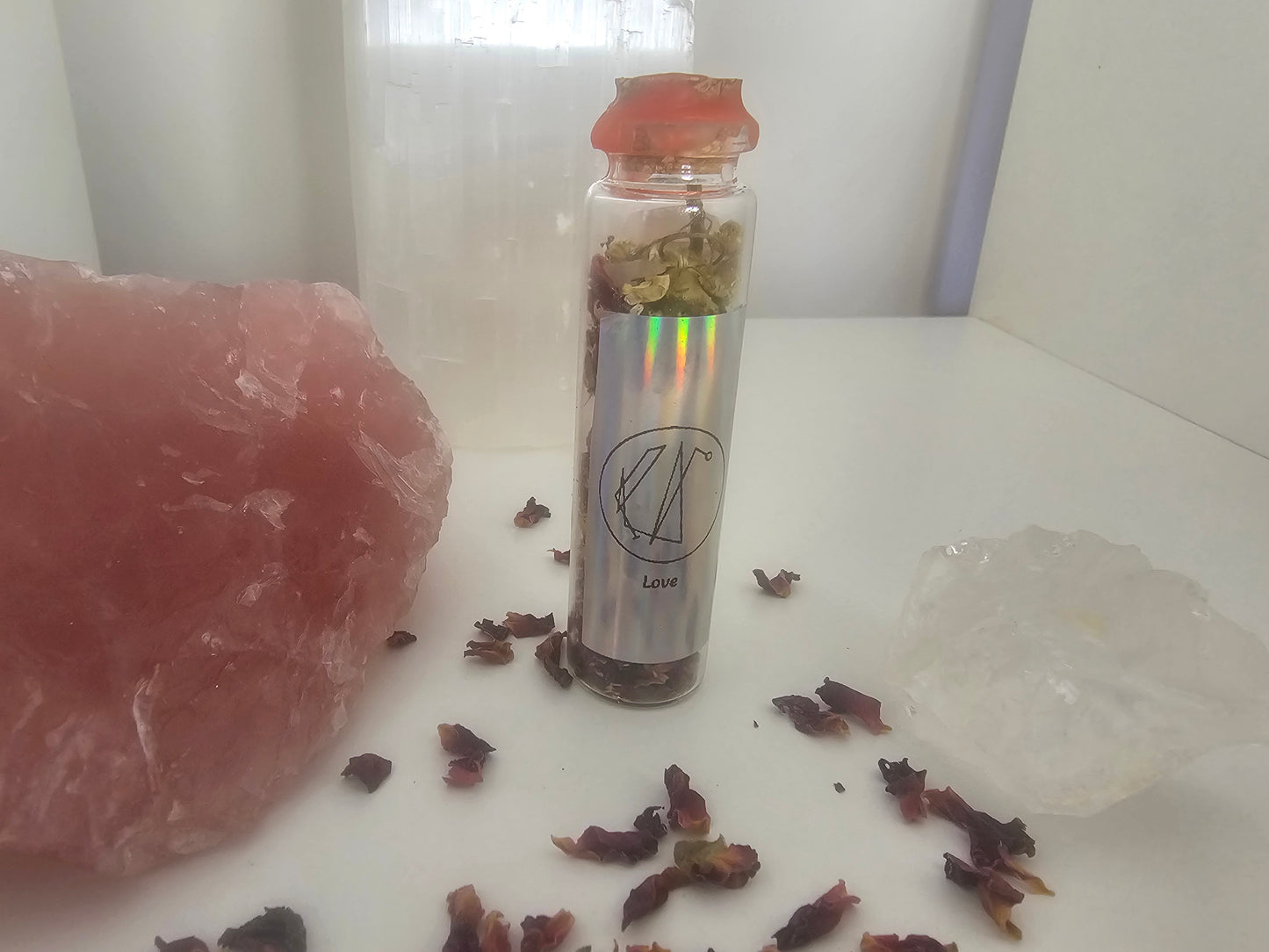 Small glass spell jar with dried herbs and crystals on a white surface. The front label has a picture sigil and it says love.