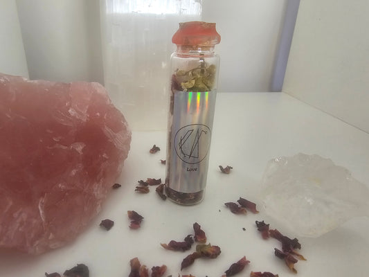 Small glass spell jar with dried herbs and crystals on a white surface. The front label has a picture sigil and it says love.