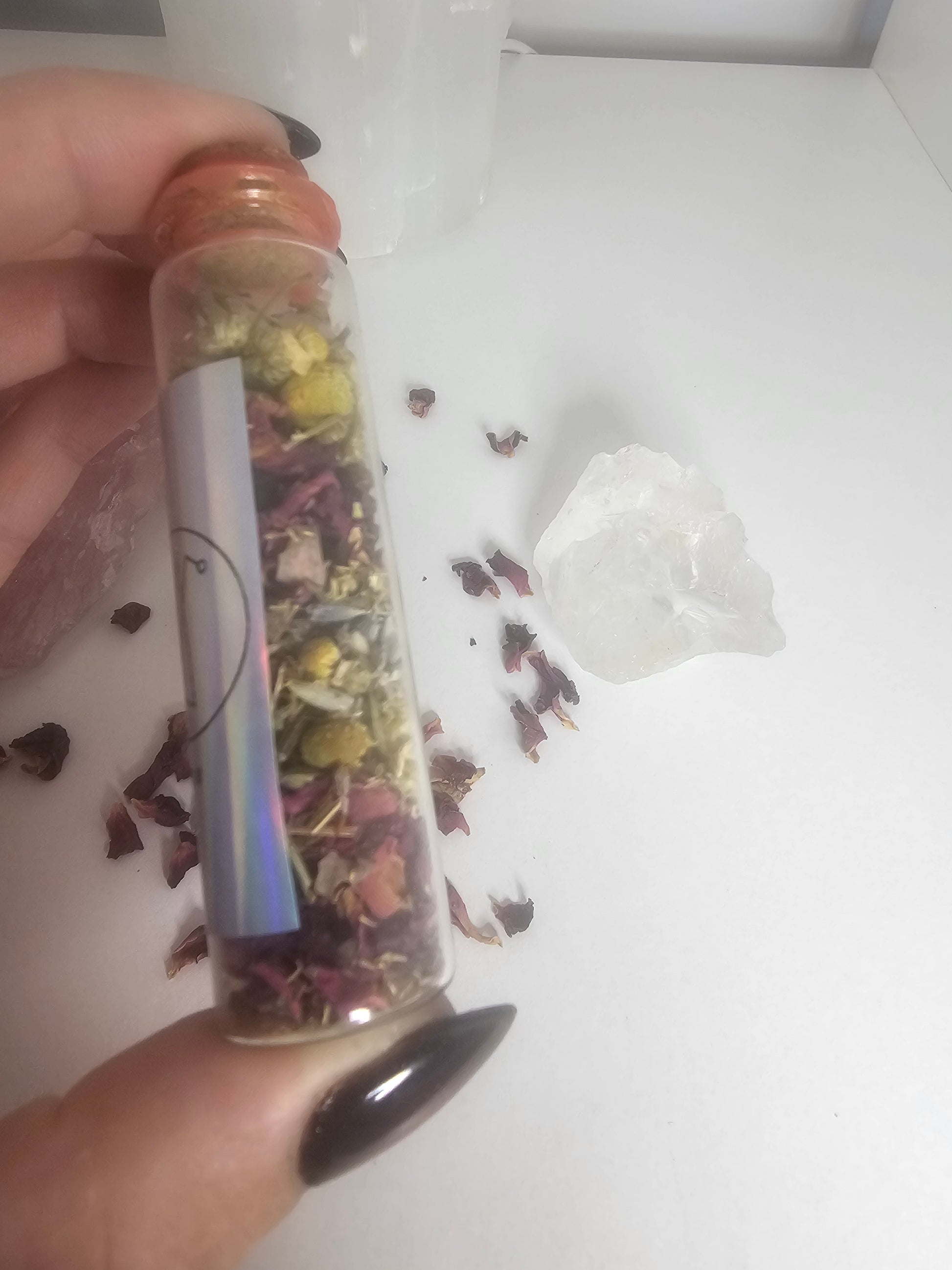 A hand with black glass nails holding a small glass spell jar filled with herbs of different colours and crystals.