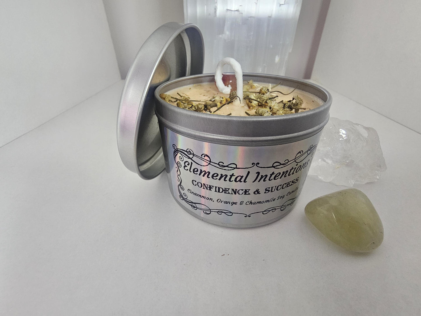 A silver candle container filled with light orange soy candle wax and topped with decorative chamomile flowers and an orange carnelian crystal. The label on the front it silver and holographic, has an ornamental black frame, and says &#39;elemental intentions, confidence and success, and cinnamon, orange and chamomile soy candle&#39;.
