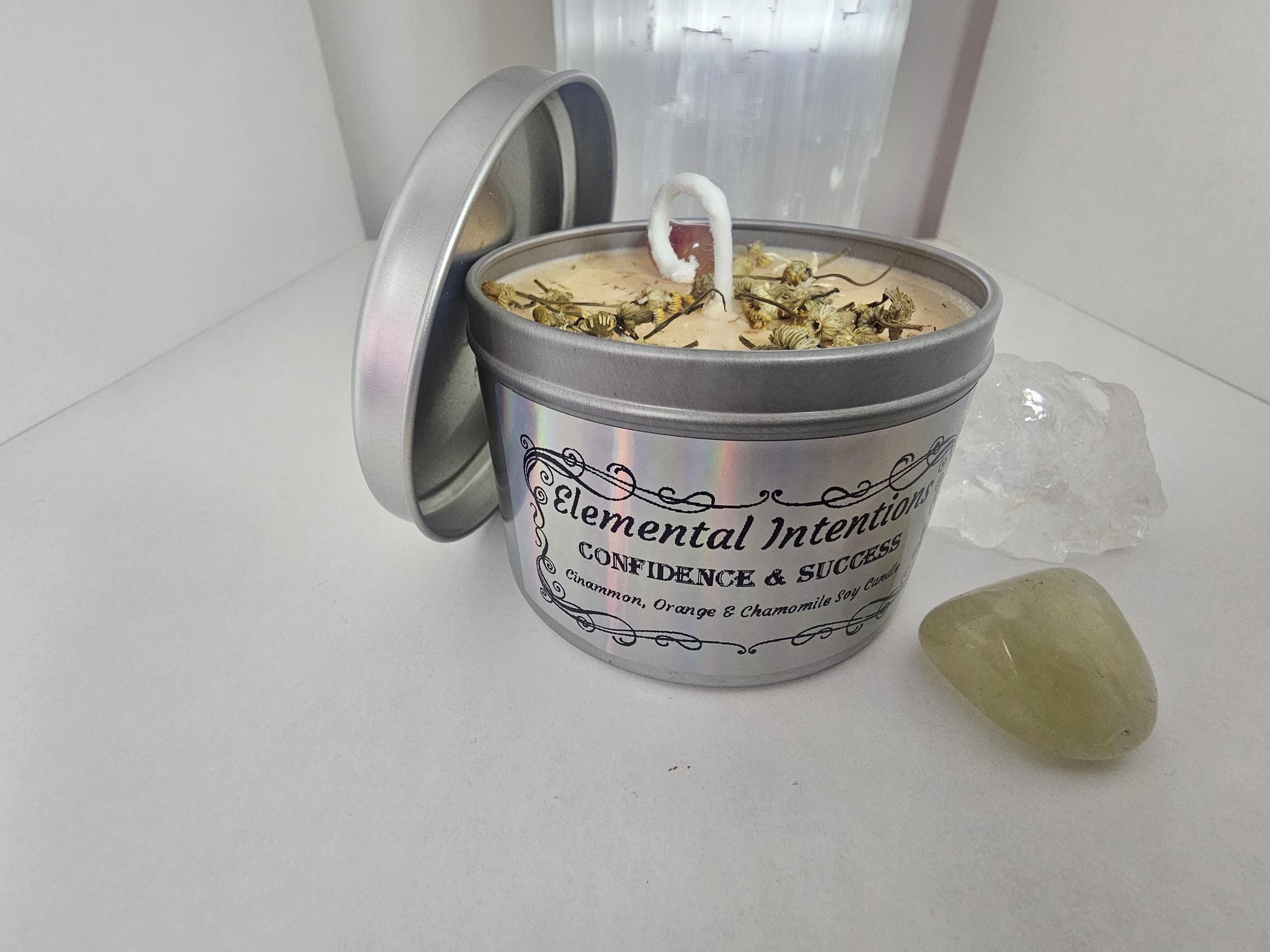A silver candle container filled with light orange soy candle wax and topped with decorative chamomile flowers and an orange carnelian crystal. The label on the front it silver and holographic, has an ornamental black frame, and says &#39;elemental intentions, confidence and success, and cinnamon, orange and chamomile soy candle&#39;.