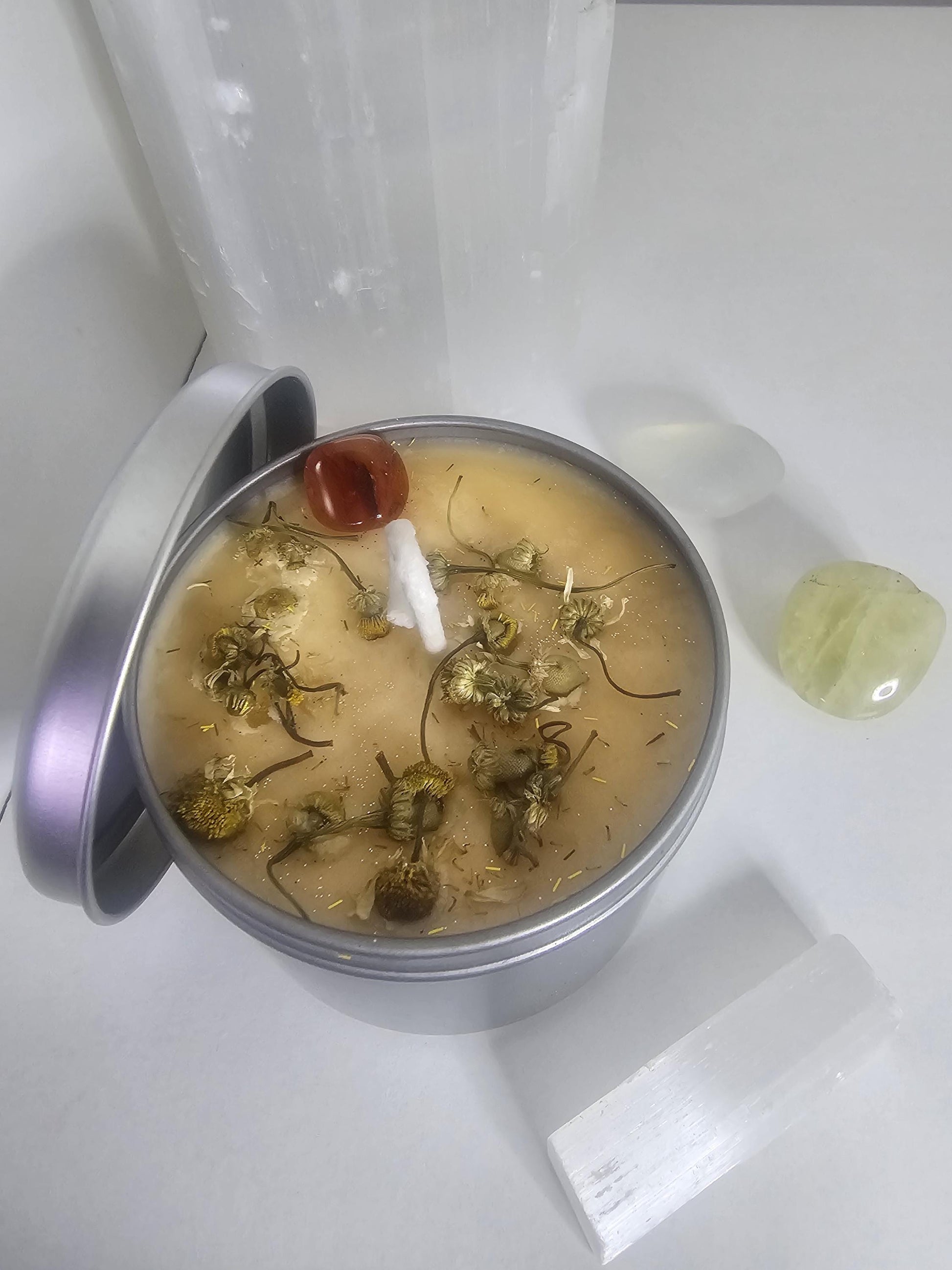 An above shot of a silver container candle with a silver matching lid resting to the side. The container is filled with light orange soy wax with a white cotton wick and is topped with decorative chamomile flowers and an orange carnelian crystal. Next to the candle lies a piece of selenite and another carnelian crystal.
