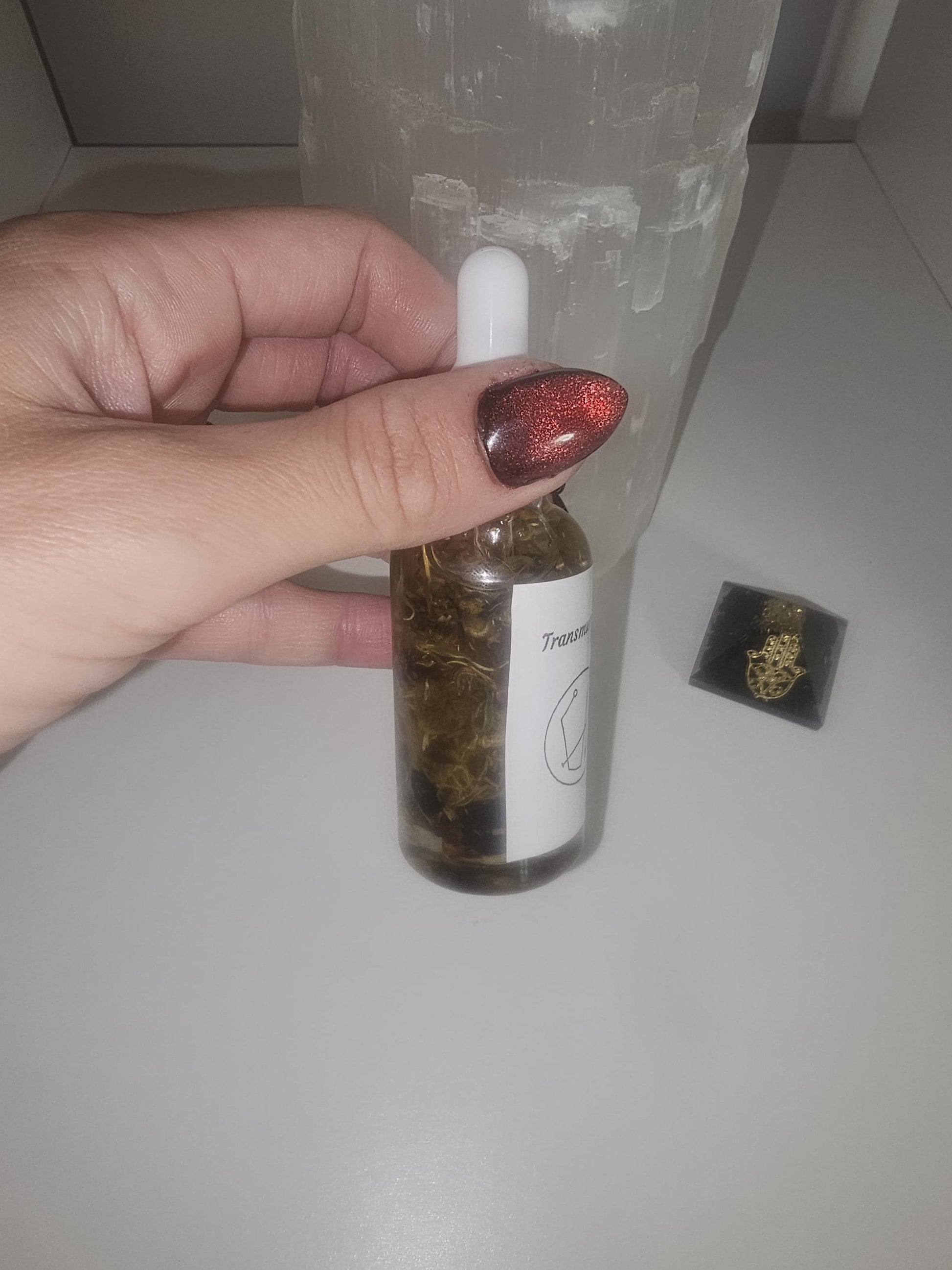 A hand with red and black glass nails holding a clear glass dropper bottle with a gold top. The bottle has a white label and is filled with oil and natural transmutation herbs.