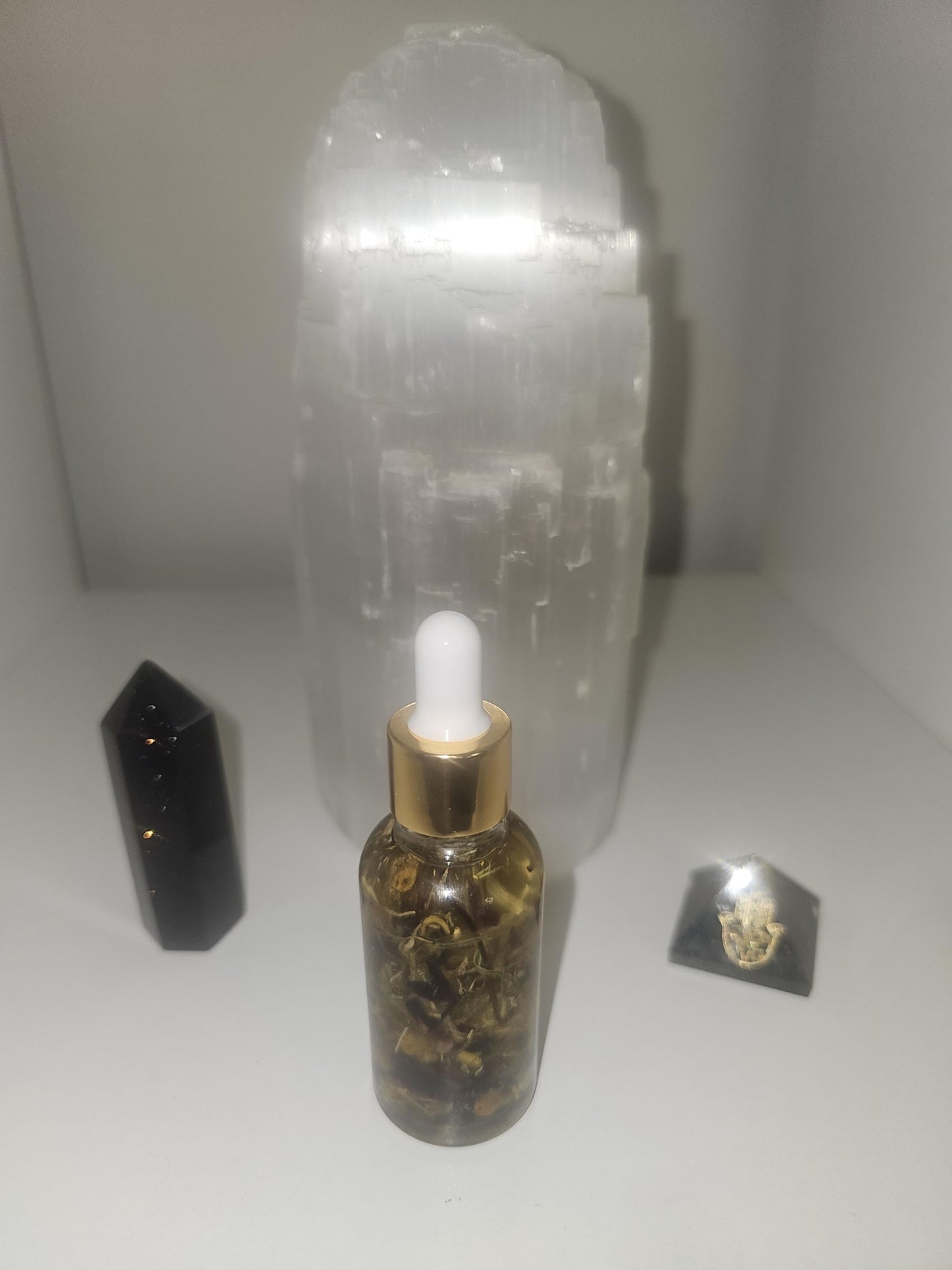 A rear sot of a glass dropper bottle filled with oil ad transmutation herbs on a white background in front of a large selenite lamp.