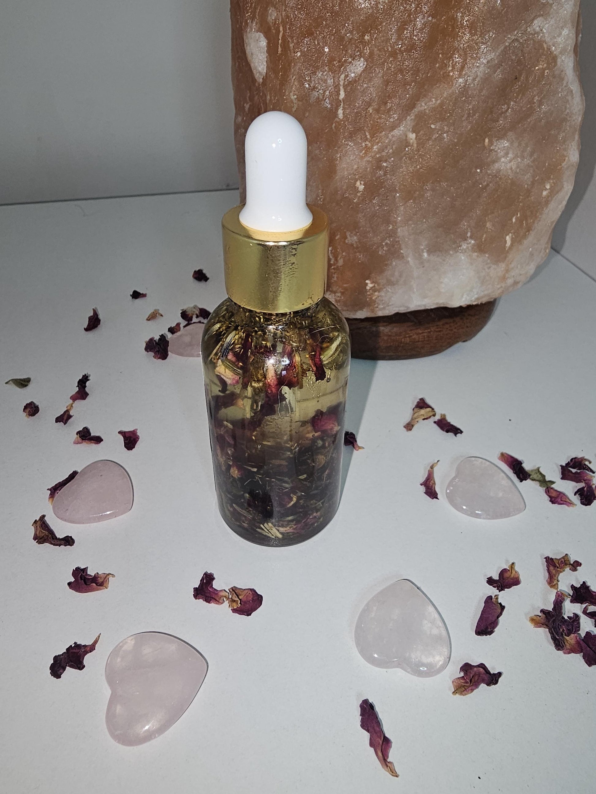 A clear glass dropper bottle with a gold top and filled with natural love and attraction herbs and oil. The bottle is surrounded by rose quartz love hears and rose petals with a rose quartz lamp by the side.