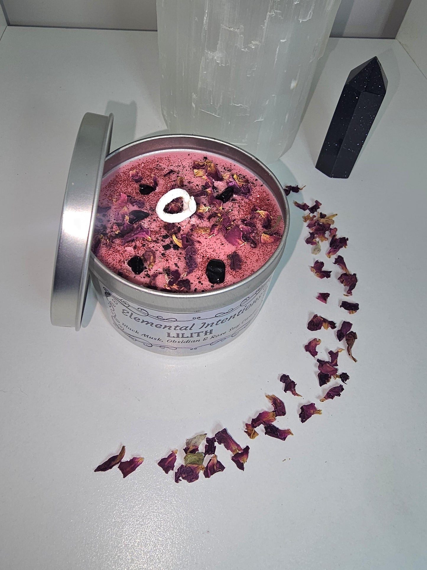 A silver candle container filled with red wax and black obsidian chips and topped with rose petals. On a white background with a moon shaped pattern of rose petals around the container and a large obsidian crystal tower to the side.