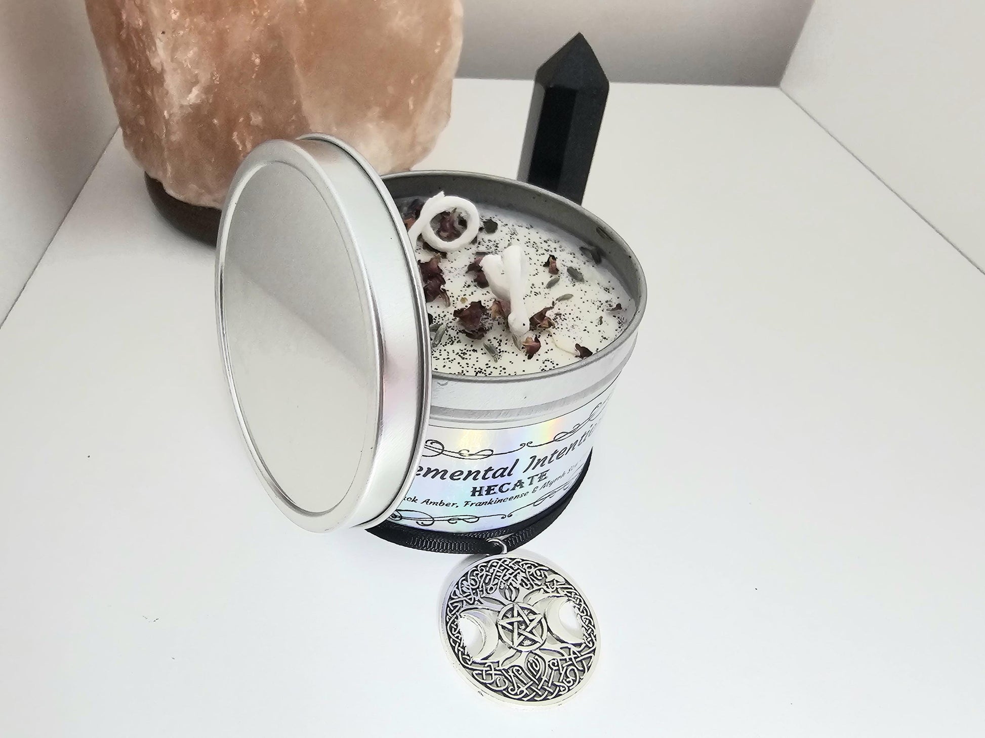 A silver container candle filled with scented white soy wax and topped with black glitter, rose petals, and with a cotton wick. The front label says &#39;Hecate&#39; and &#39;elemental intentions&#39; and there is a black necklace with a triple moon pendant surrounding the candle on a white background.
