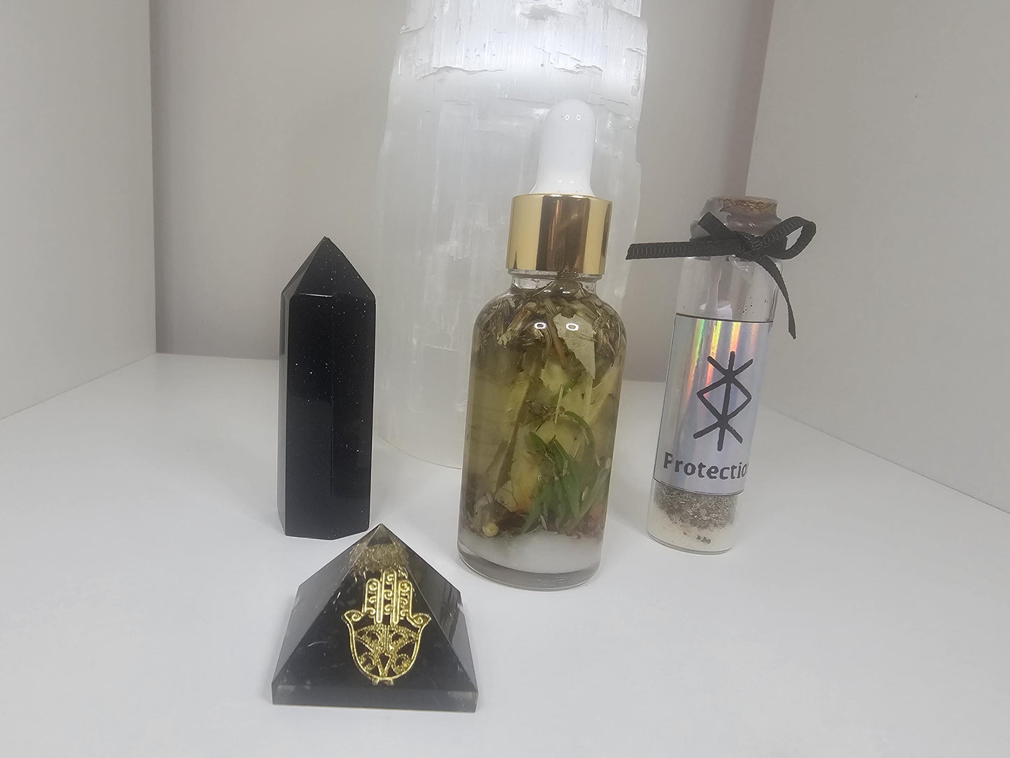 A shot of a clear glass dropper bottle with a gold lid filled with natural protection herbs. Stands next to a black and gold hamza pyramid, a black obsidian tower and a large selenite lamp.