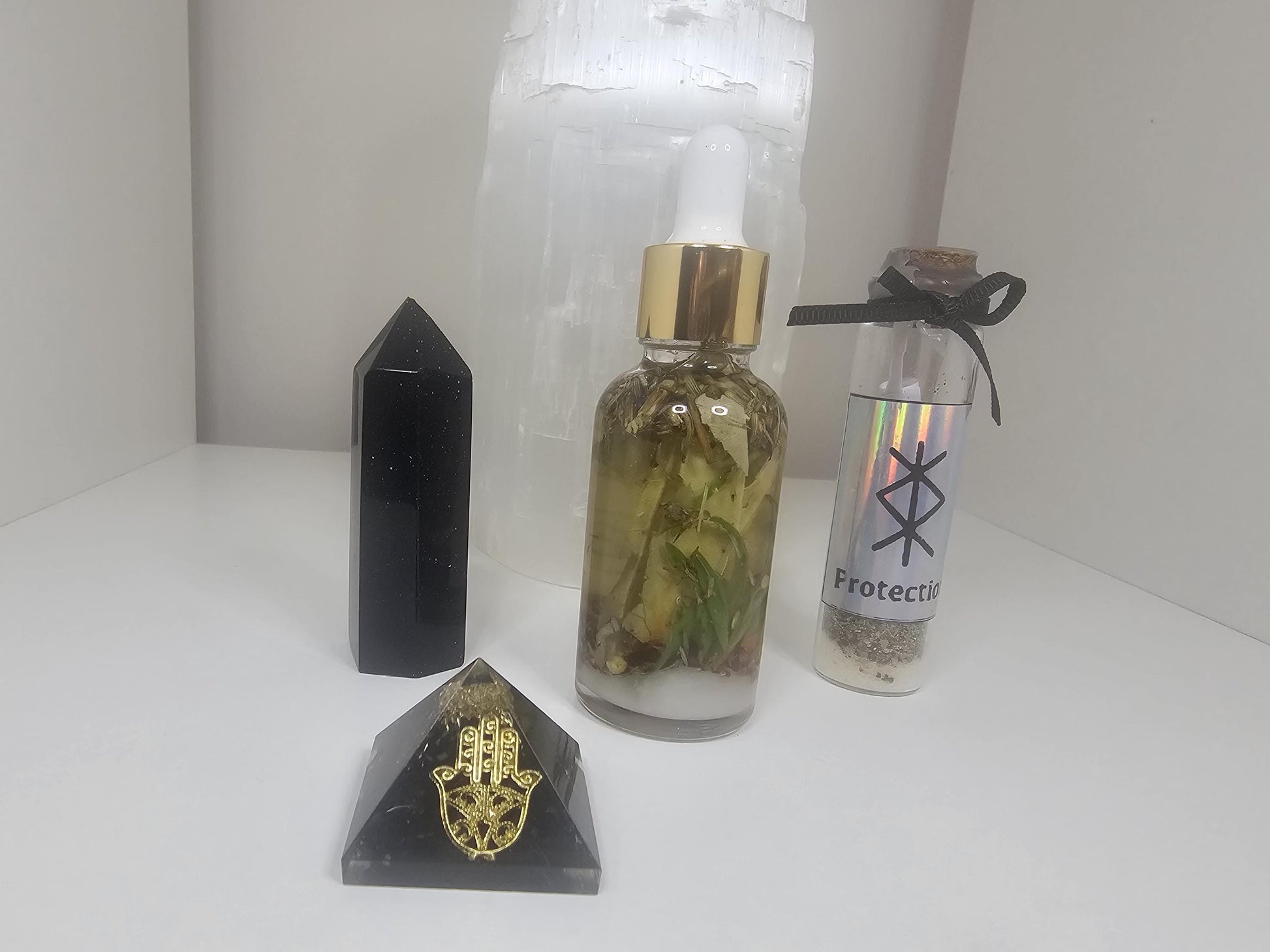 A shot of a clear glass dropper bottle with a gold lid filled with natural protection herbs. Stands next to a black and gold hamza pyramid, a black obsidian tower and a large selenite lamp.