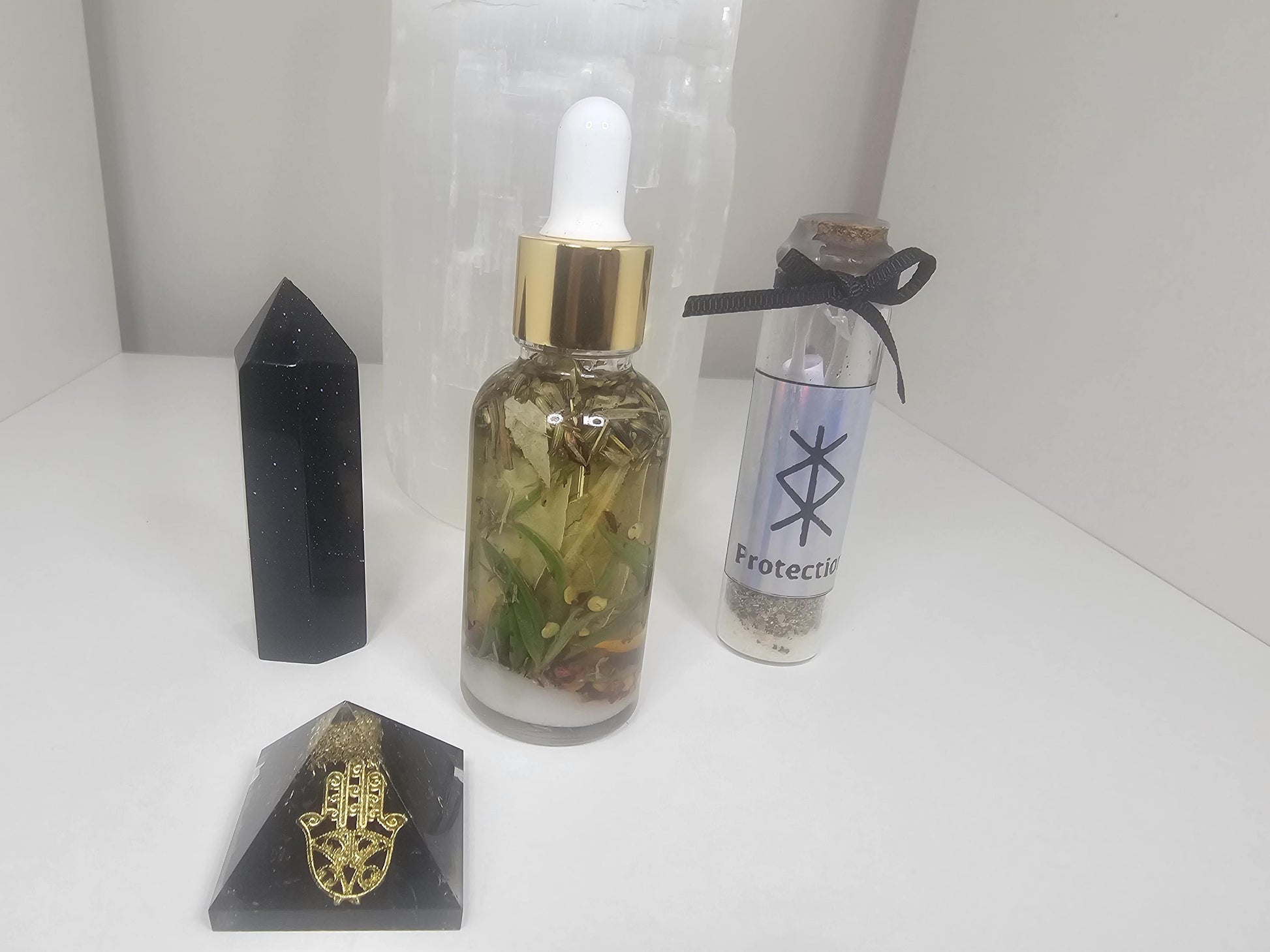 A 30ml clear dropper bottle with a gold lid, filled with protection herbs. The bottle stands next to a selenite lamp, a black obsidian tower and a protection spell jar.