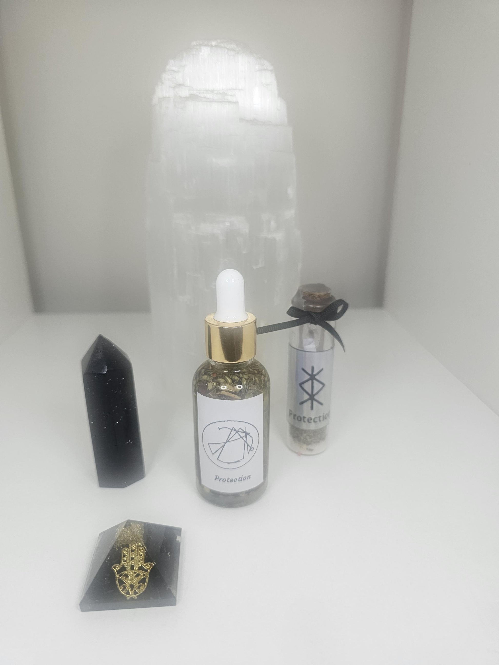 A front shot of a clear glass dropper bottle with a gold lid and a white label with a protection sigil on the front. The bottle is filled with natural protective herbs and stands next to a gold and black hamza pyramid, a black obsidian tower and a large selenite lamp.