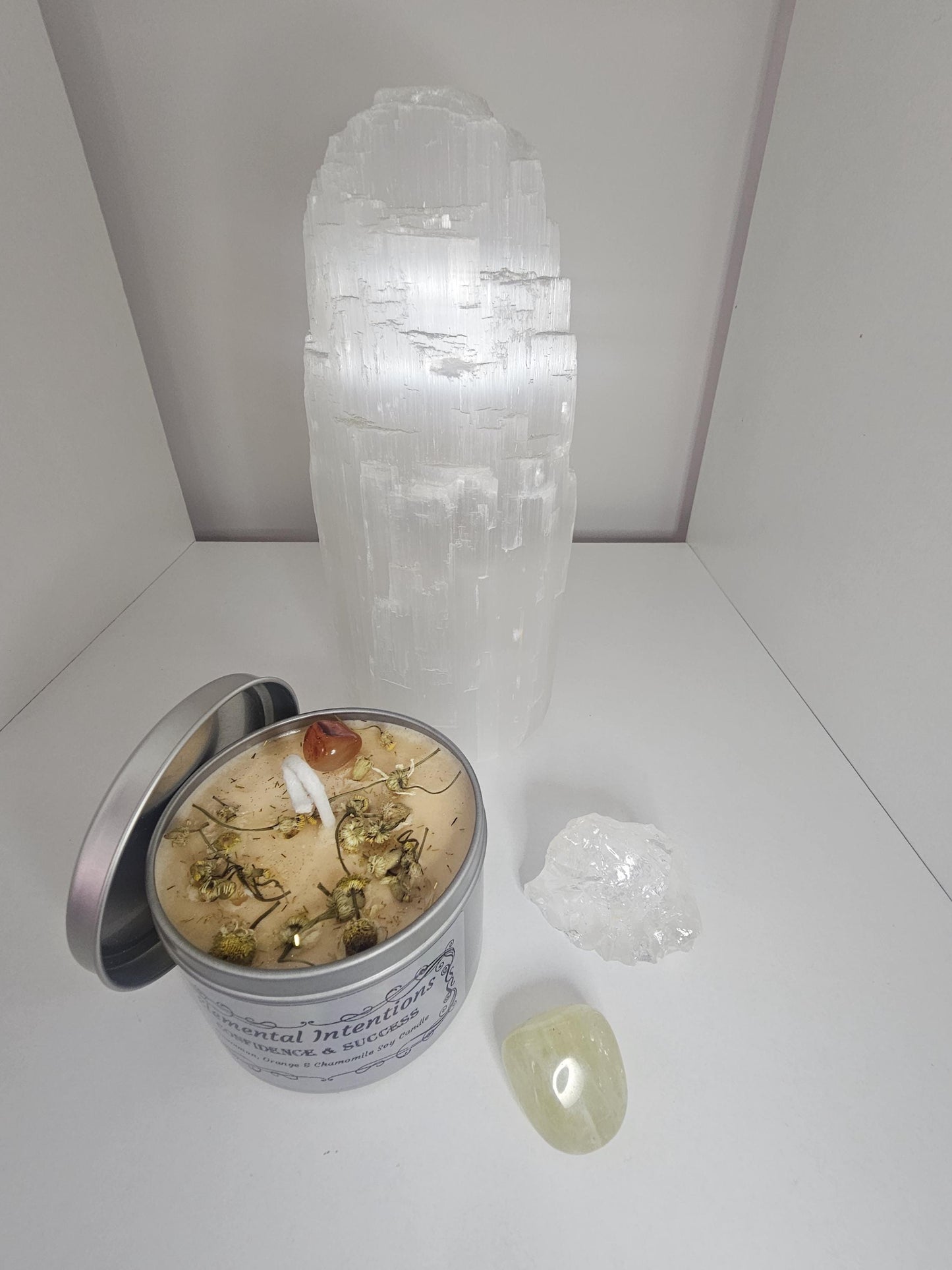 A light orange soy candle in front of a large selenite crystal lamp, and in a silver container. The candle is topped with chamomile flowers and an orange carnelian crystal.