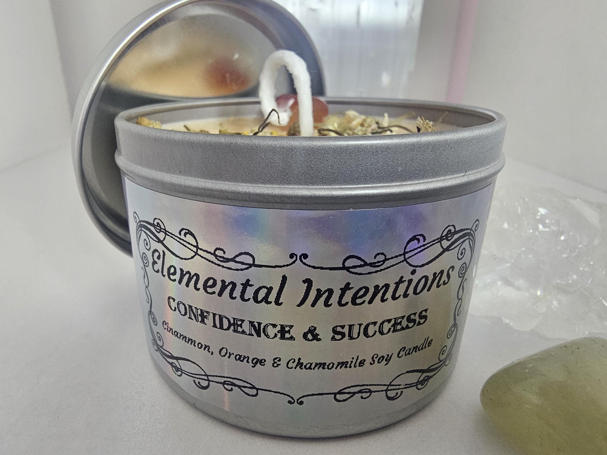 A front shot of a silver container candle with a holographic silver label which has black ornamental framing. The words read &#39;elemental intentions, confidence and success, and cinnamon, orange and chamomile soy candle&#39;.