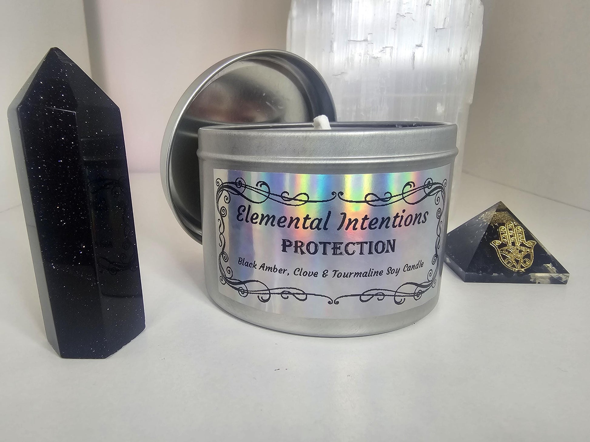 A front shot of a silver container candle with a holographic label containing ornate black framing and the words &#39;elemental intentions, protection, and Black Amber, Clove and Tourmaline soy candle&#39;