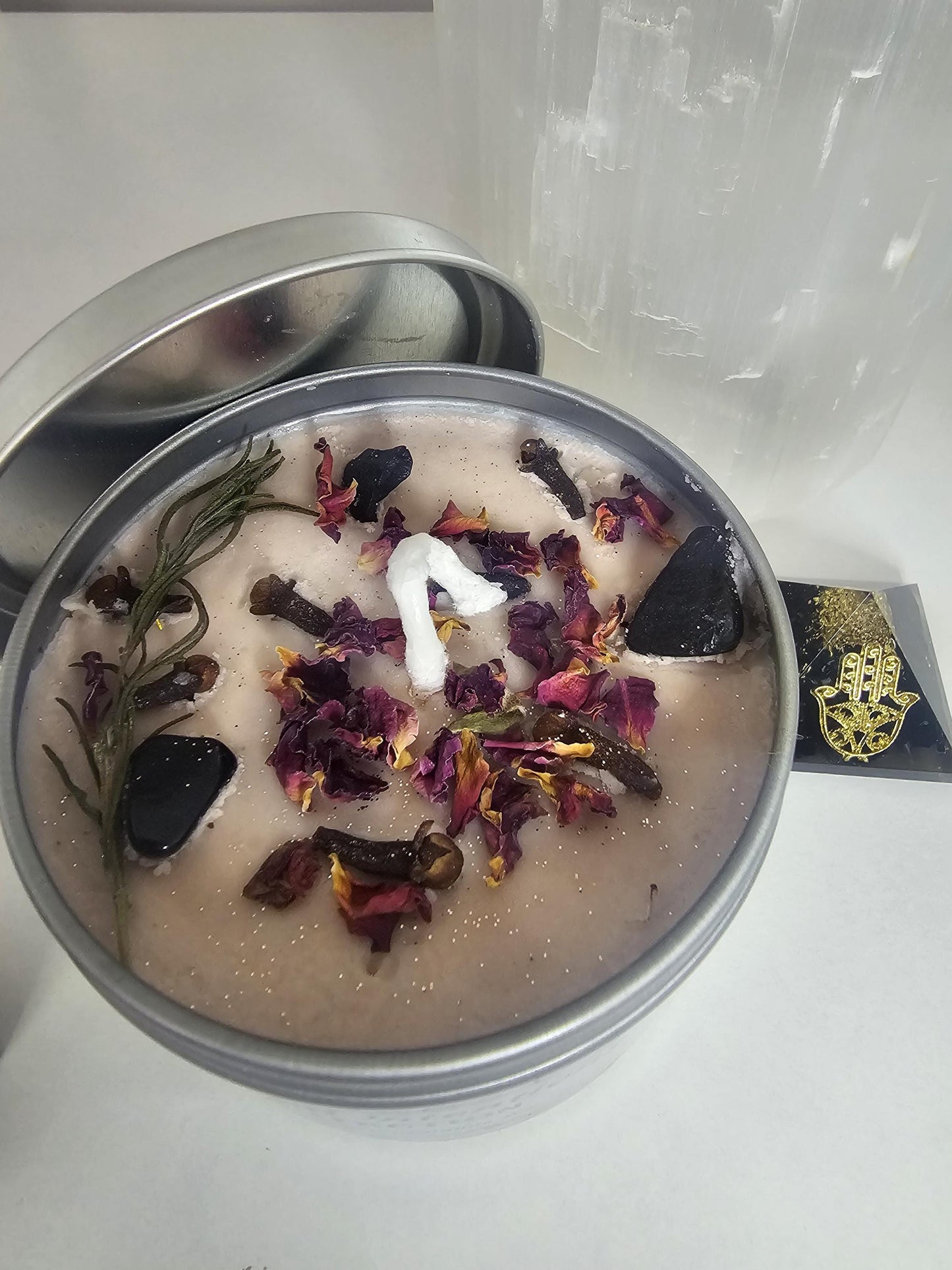 A close up of a nude coloured wax in a silver candle container, topped with shimmering rose gold glitter, rose petals, and black tourmaline crystals.