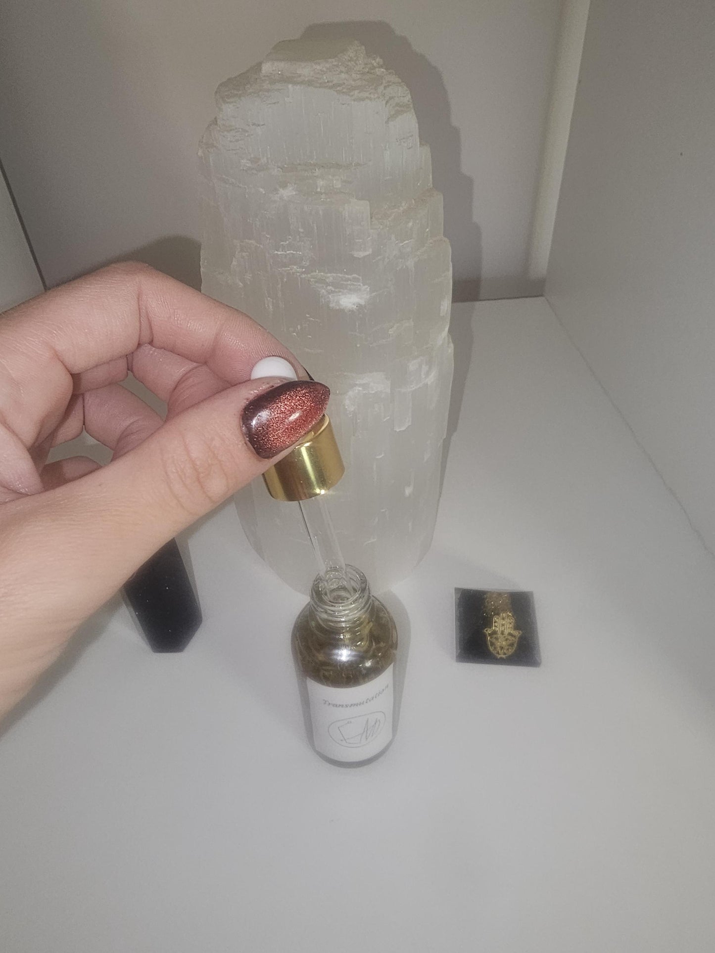 A hand with red and black glass nails holding a god dropper top of a clear glass bottle, fille with oil and transmutation herbs on a white background.