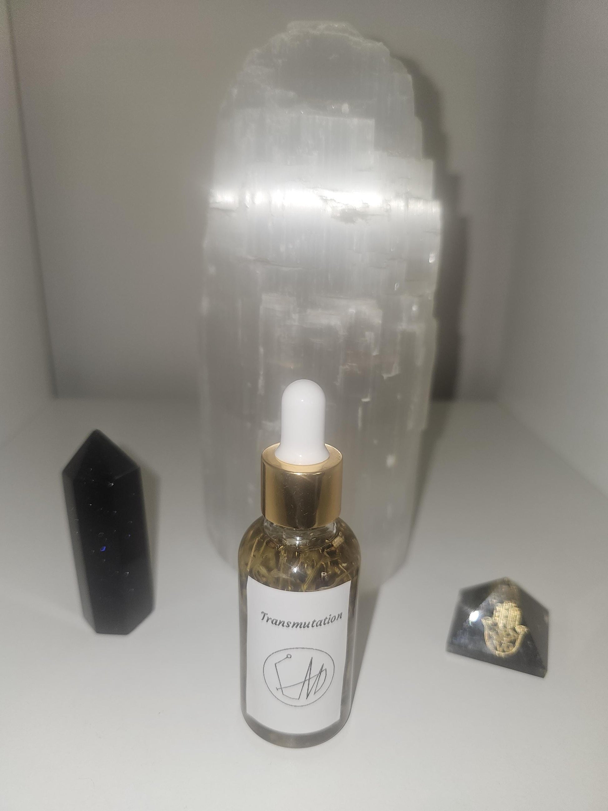 A clear glass dropper bottle with a gold top filled with oil and natural transmutation herbs. The bottle is in front a large selenite crystal lamp and surrounded by a gold and black hamza pyramid and a black tourmaline crystal tower.