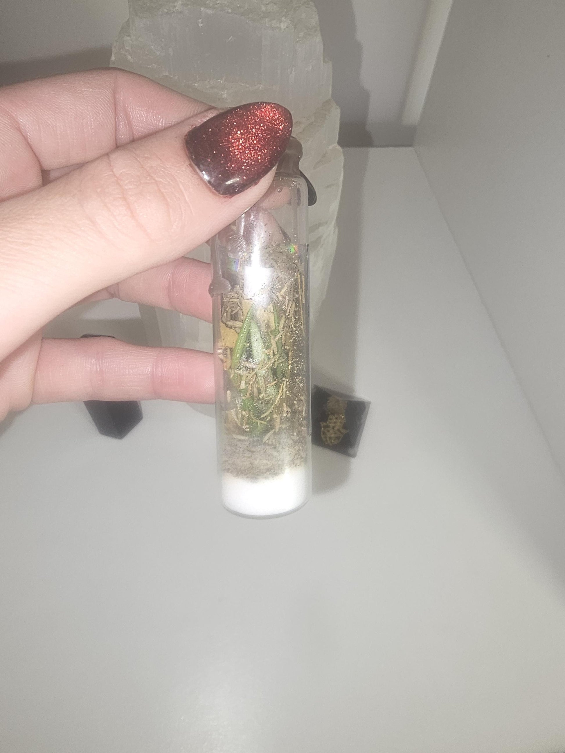 A hand with black and red glass nails holding a small glass jar that is filled with protective herbs and a personalised scroll, which has been sealed with black wax.