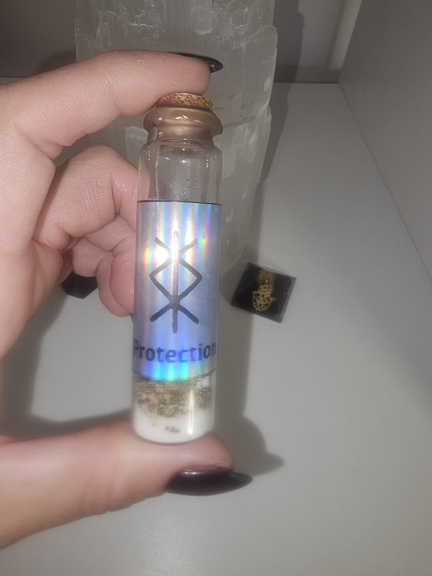 A hand with red and black glass nails, holding a small glass jar with a silver holographic label on the front containing the word &#39;protection&#39; and a protection sigil.