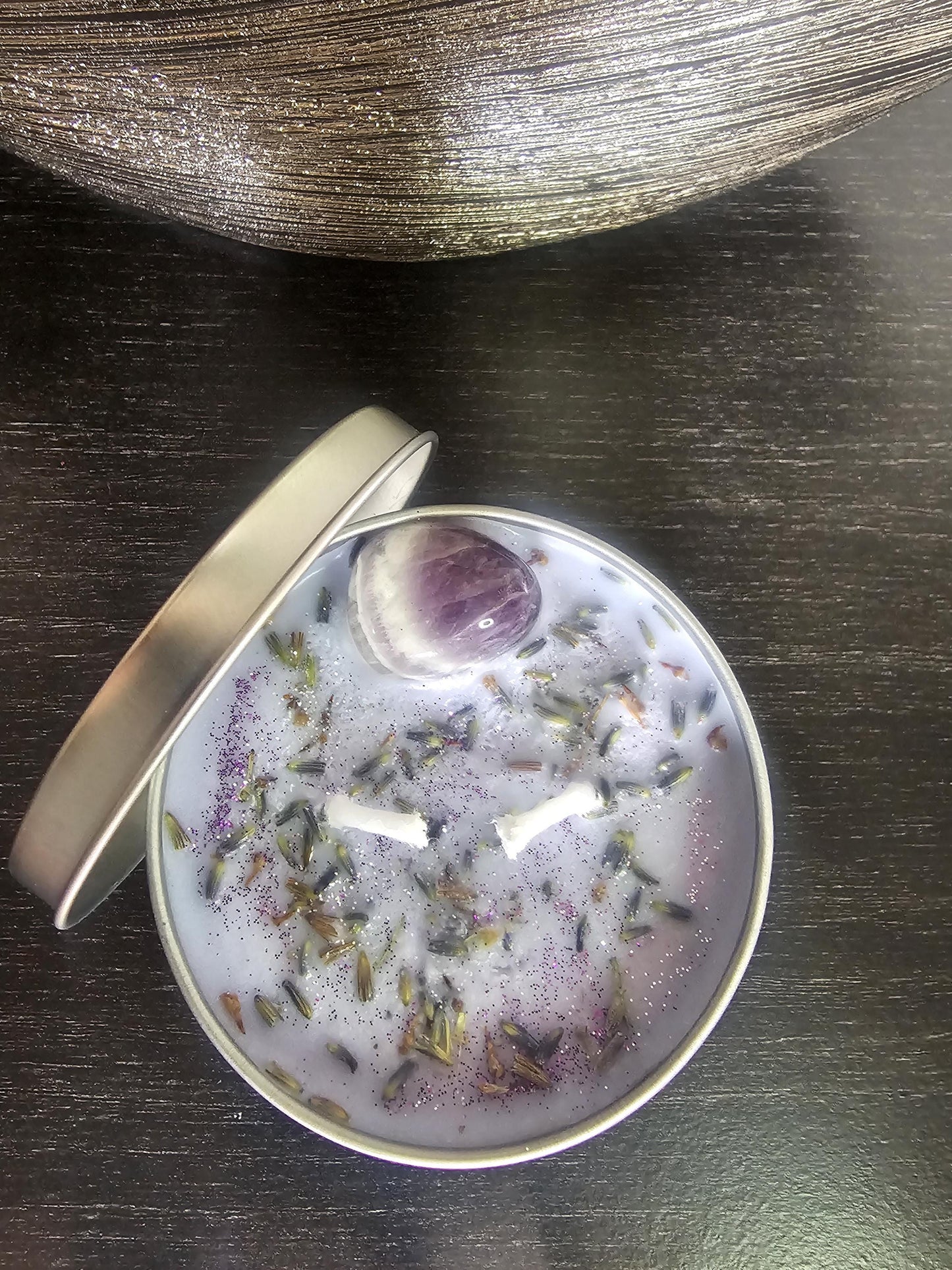 An above shot of a silver candle container on a black background, filled with light purple soy wax and topped with decorative lavender flowers and a medium sized amethyst crystal.