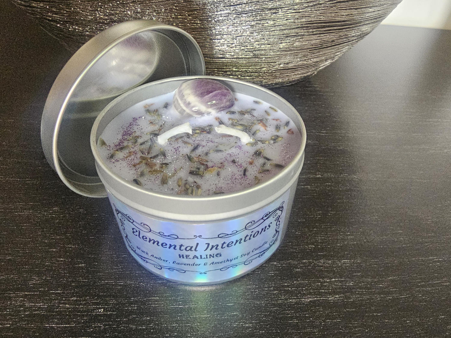 A silver candle container on a black background filled with light purple wax and topped wih lavender flowers, purple glitter and a purple amethyst crystal.