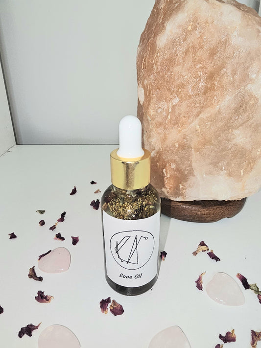 A clear glass dropper bottle with a gold top and a white label saying &#39;love&#39; and containing a love and attraction sigil. Th bottle is filled with natural attraction and love herbs and is surrounded by a rose quartz lamp and rose petals.