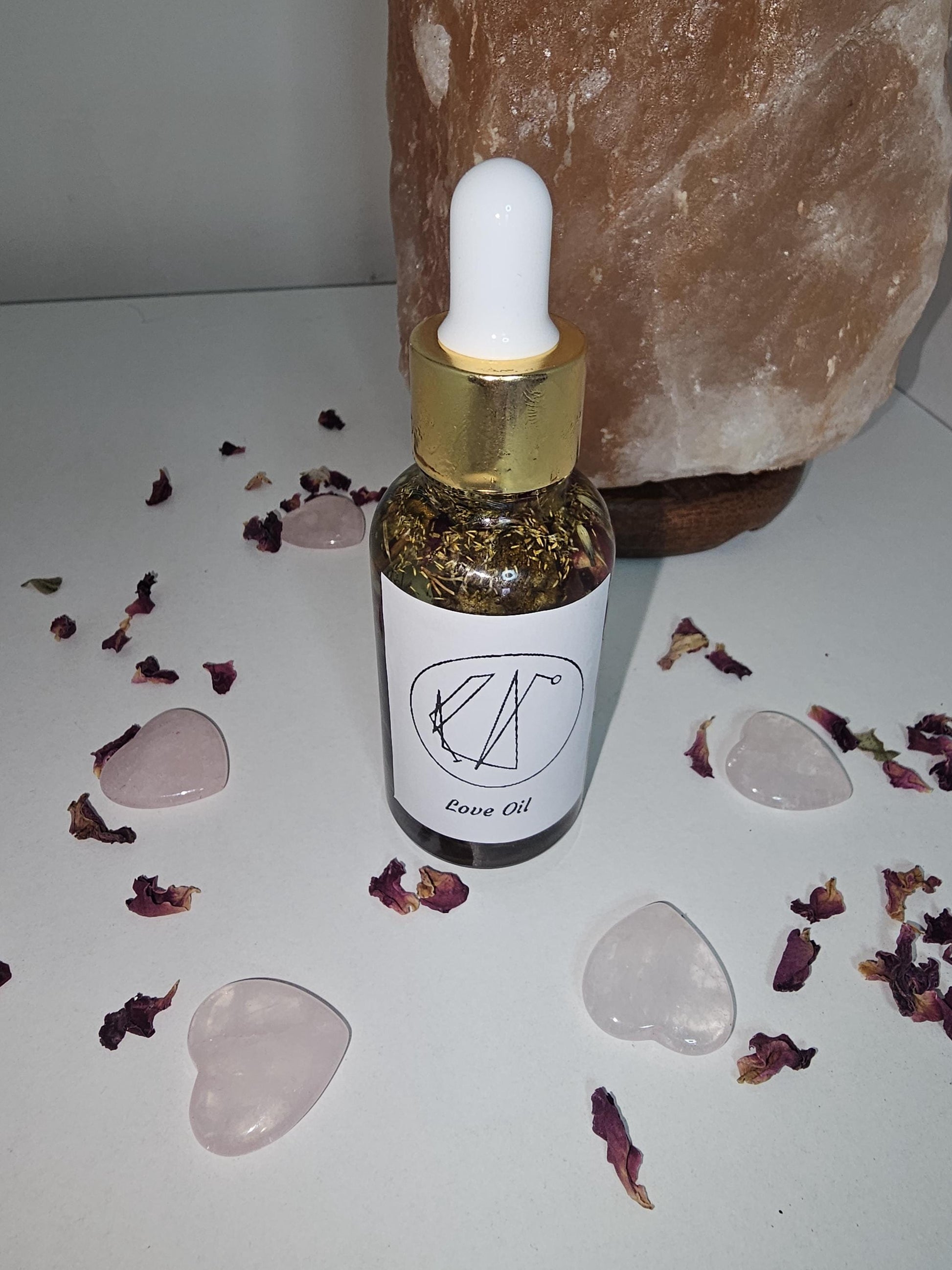 A close up of a clear glass dropper bottle with a gold top filled with herbs and oil and surrounded by rose petals, rose quartz love hearts and a large rose quartz lamp to the side.
