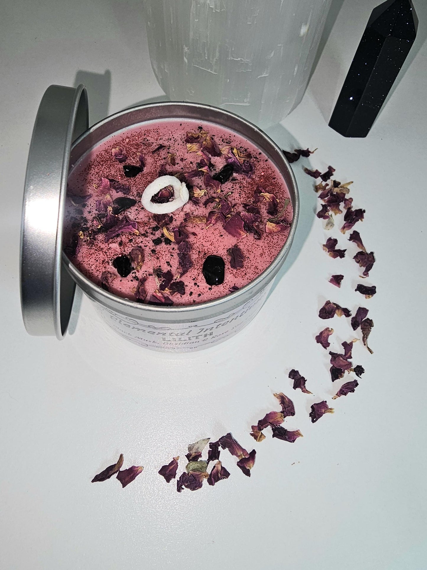 An above shot of a silver container candle filled with red wax and topped with rose petals and obsidian crystals, with a cotton wick.