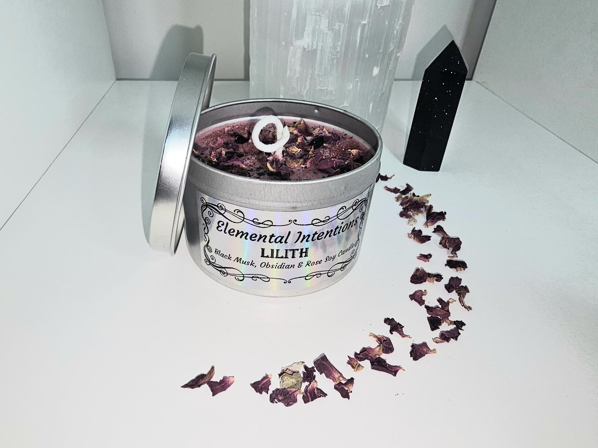 A front shot of a silver candle holder filled with red wax and topped with rose petals and black obsidian chips. The front label is framed with ornate black framing and the words read &#39;elemental intentions&#39;, &#39;Lilith&#39; and the title &#39;Black musk, rose and obsidian soy candle&#39;.