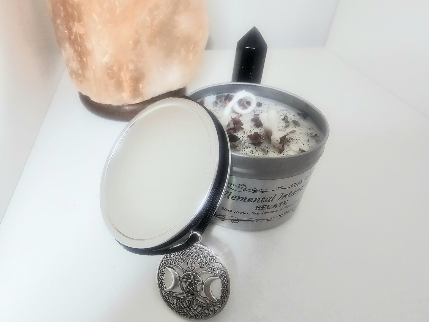 A silver container candle with a silver lid placed next to it surrounded by a black necklace with a triple moon pendant. The candle is white and topped with rose petals, black tourmaline, and black glitter.