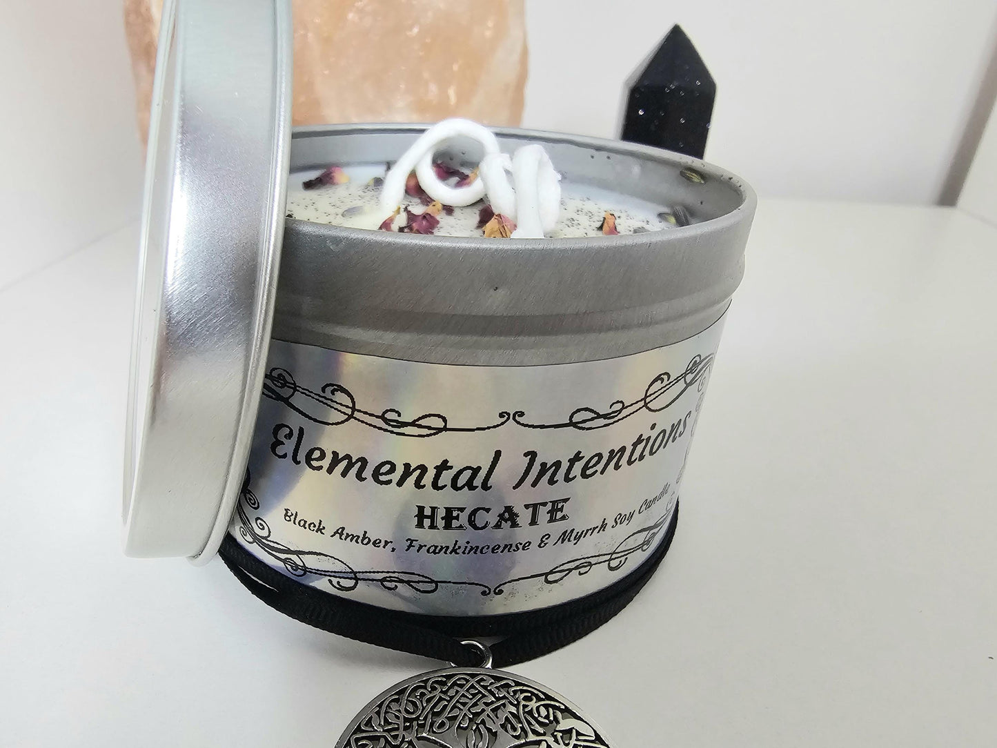 A front close up of a silver container candle with an ornamental black frame on a holographic label. The label is framed with a black ornate frame and says &#39;elemental intentions&#39;, &#39;Hecate&#39; and &#39;Black Amber, Frankincense and Myrrh soy candle&#39;.