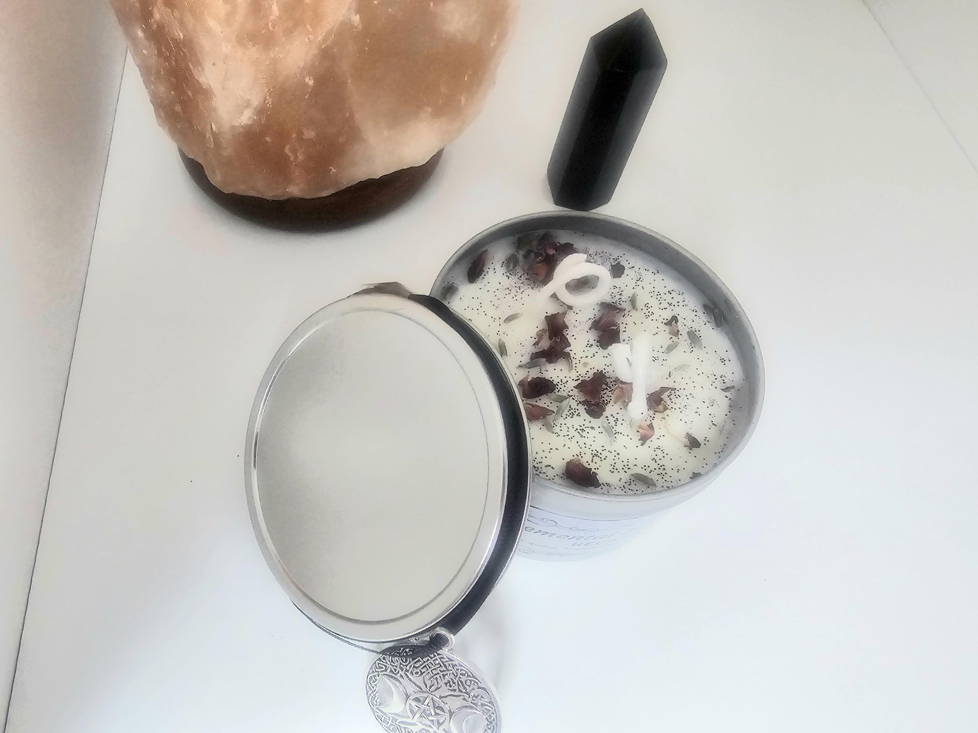 An above shot of a white scented candle in a silver container surrounded with a black necklace with a triple moon pendant. The candle has white cotton wicks and is topped with black tourmaline crystals, rose petals and black glitter.