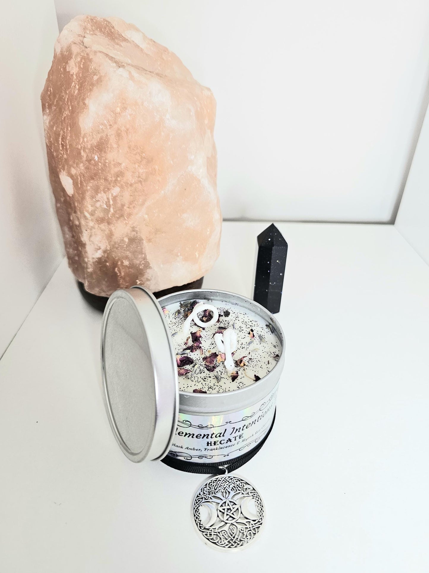 An above shot of a white scented candle in a silver container, topped with black tourmaline, rose petals and black glitter. The candle is surrounded with a triple moon pendant and a black tourmaline crystal tower.