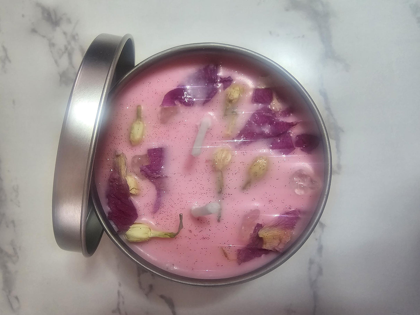 An above shot of a silver candle container filled with light pink soy wax on a marble table. the candle is decorated with pink lotus petals and white jasmine buds, and shimmers with pink and silver glitter.