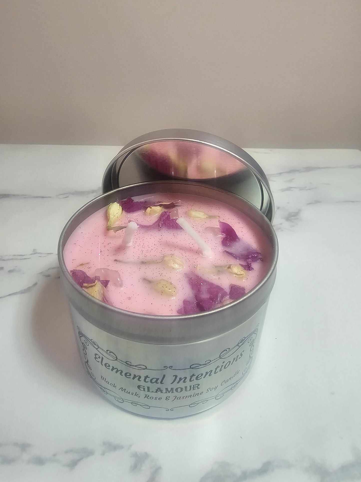 A silver candle container with light pink wax and decorative jasmine buds and lotus petals with two white cotton wicks. A lid rests behind the candle container on a marble table.