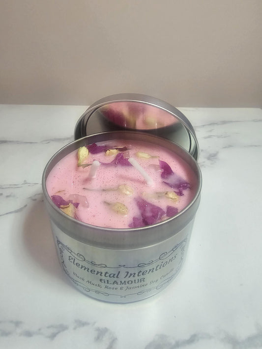 A silver candle container with light pink wax and decorative jasmine buds and lotus petals with two white cotton wicks. A lid rests behind the candle container on a marble table.