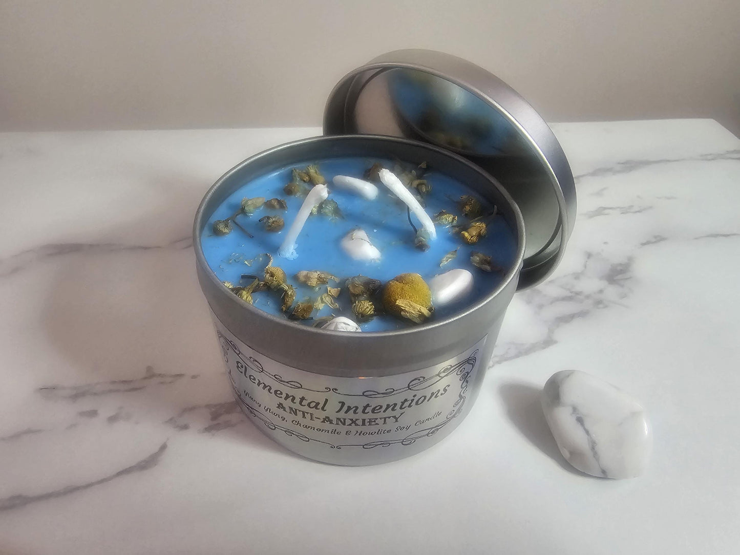 An above shot of a silver container aromatherapy candle in blue topped with chamomile decorative pieces and small howlite crystals to reduce anxiety. There is an ornamental label slightly in view and it is sat on a white marble table next to a larger howlite crystal.