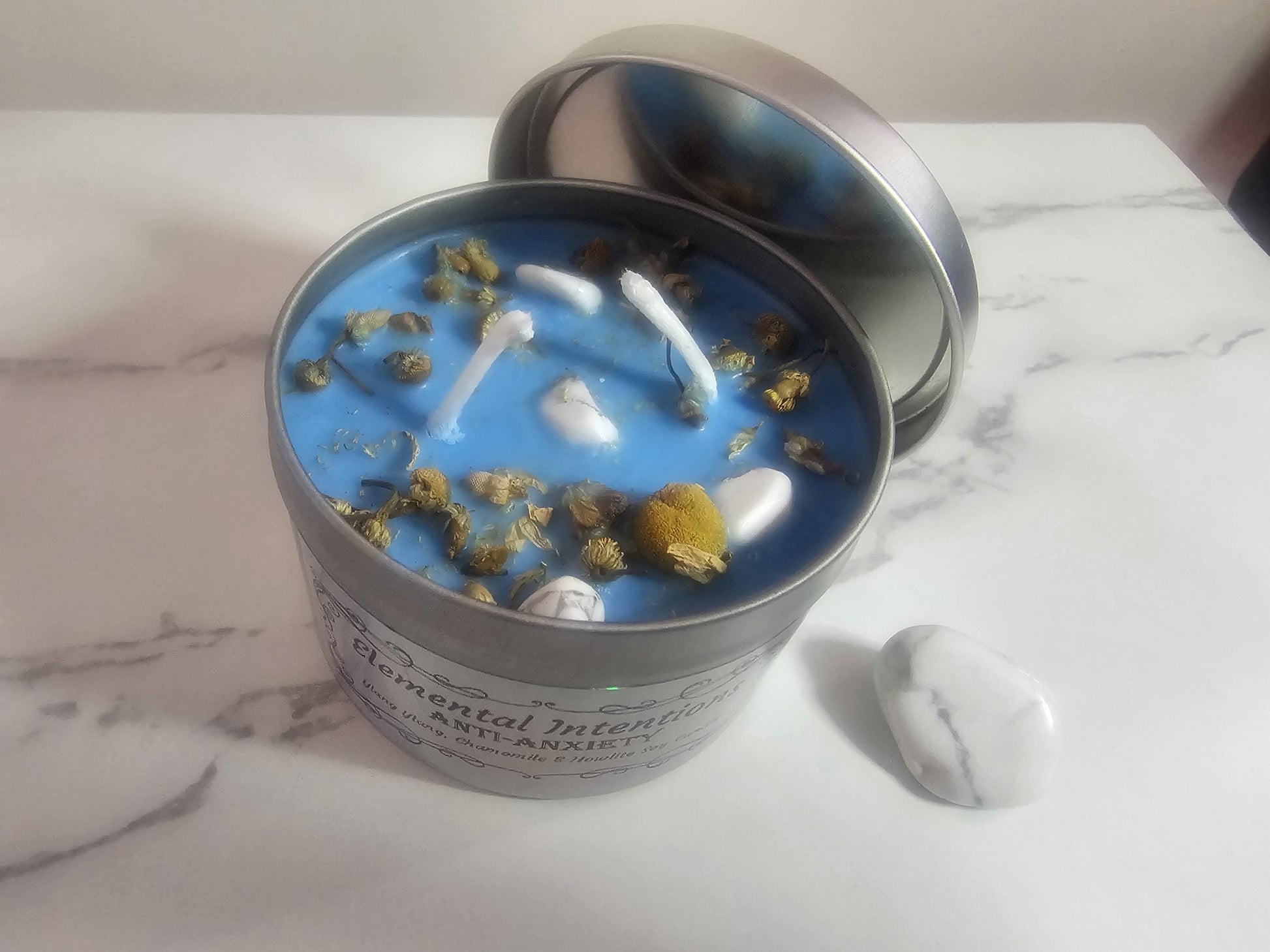 An above shot of a silver container aromatherapy candle in blue topped with chamomile decorative pieces and small howlite crystals to reduce anxiety. There is an ornamental label slightly in view and it is sat on a white marble table next to a larger howlite crystal.