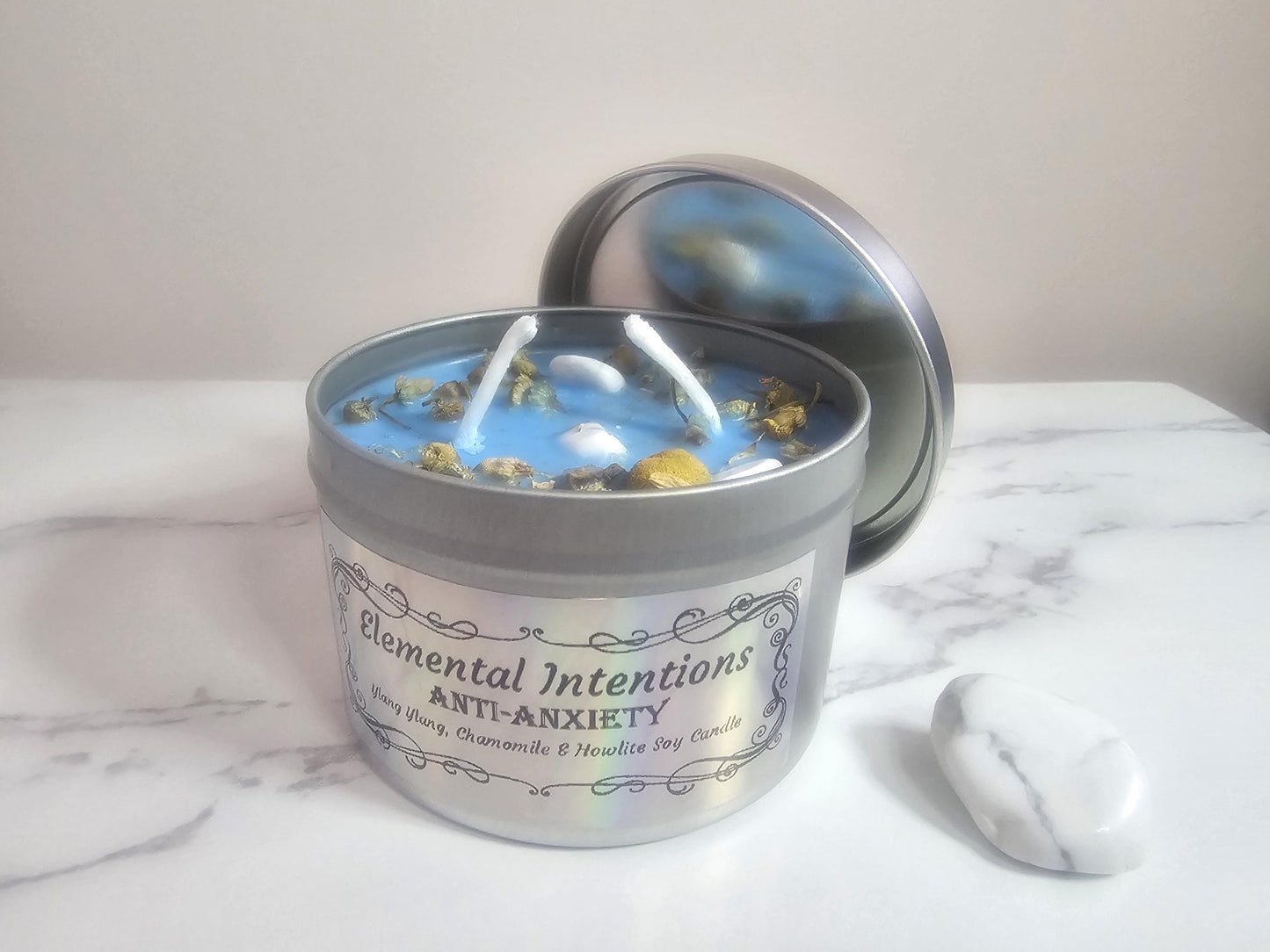 A front shot of a silver container aromatherapy candle in light blue on a white marble background next to a white howlite crystal. The candle has two wicks and is topped with chamomile and small howlite crystals. The label is framed with an oriental fine lined frame with the words &#39;Elemental Intentions, &#39;anti-anxiety&#39; and ylang ylang, chamomile and howlite soy candle&#39; on the front.