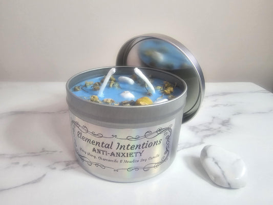 A front shot of a silver container aromatherapy candle in light blue on a white marble background next to a white howlite crystal. The candle has two wicks and is topped with chamomile and small howlite crystals. The label is framed with an oriental fine lined frame with the words &#39;Elemental Intentions, &#39;anti-anxiety&#39; and ylang ylang, chamomile and howlite soy candle&#39; on the front.