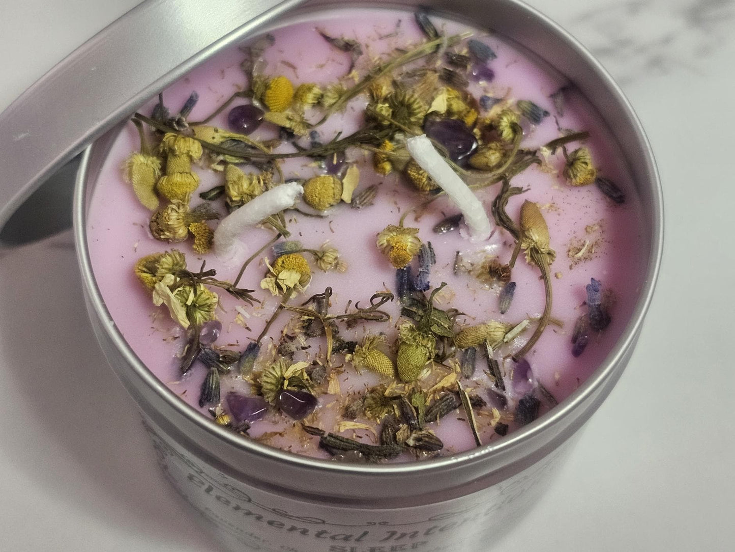 An above shot of a silver container candle which is light purple in hue and topped with decorative chamomile pieces and small amethyst crystal chips.