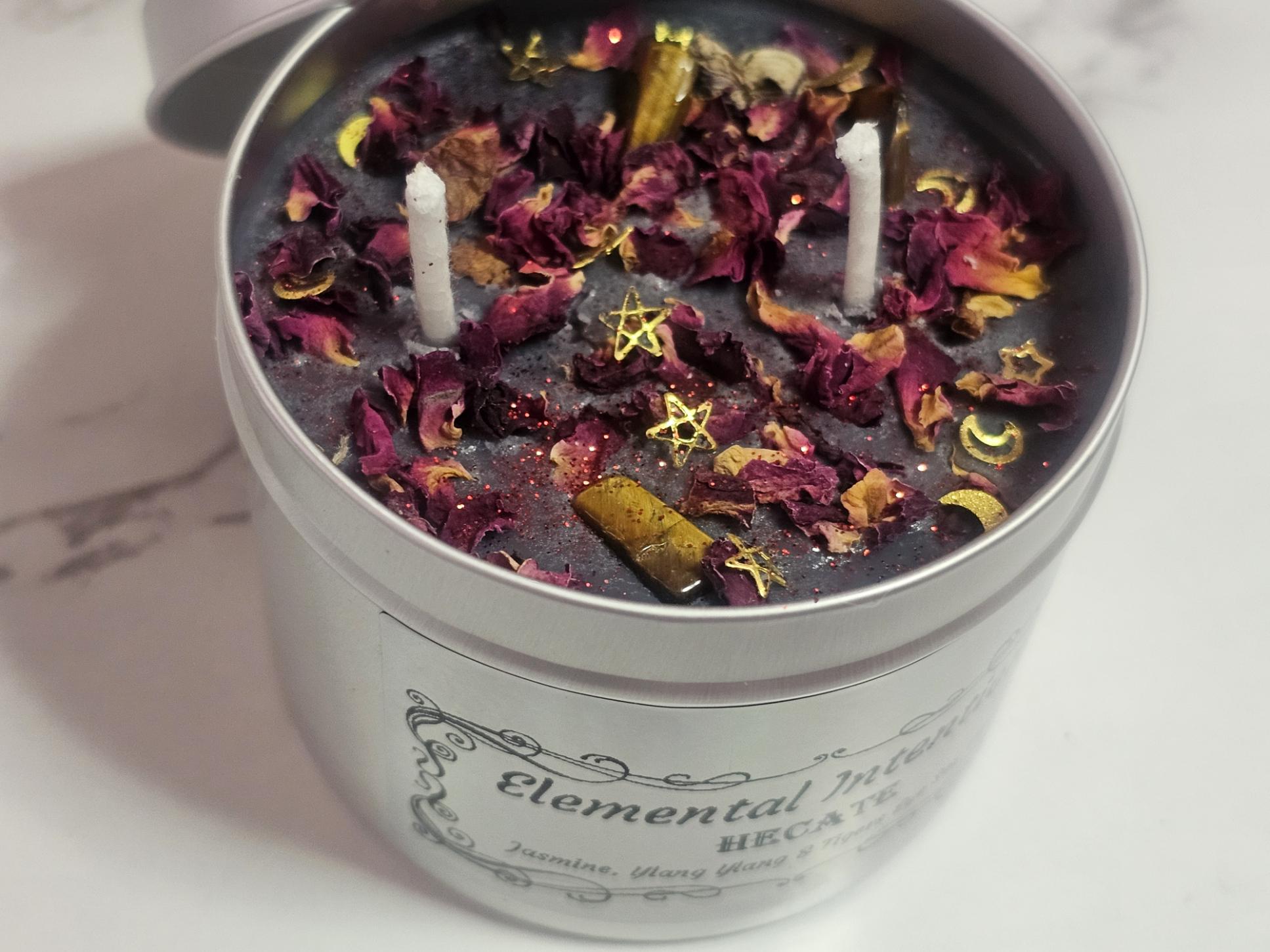 A silver container candle with a luminescent silver label saying &#39;Elemental Intentions, and Hecate&#39;. Label states the scent is Jasmine, Ylang Ylang and it has tigers eye crystals as decorative toppers with rose petals and gold moon and stars decorative pieces along with two white cotton wicks. There is a silver lid to the back which goes on the top and it&#39;s on a white marble background.